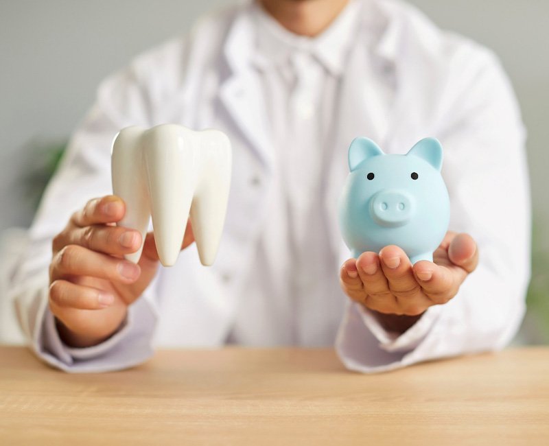 Dentist in white coat holding model tooth and blue piggy bank