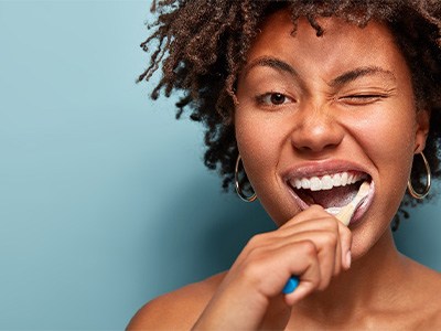 Woman with brown hair and gold hoop earrings brushing her teeth winking