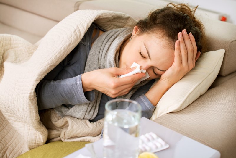 A sick woman lying on a couch and blowing her nose.