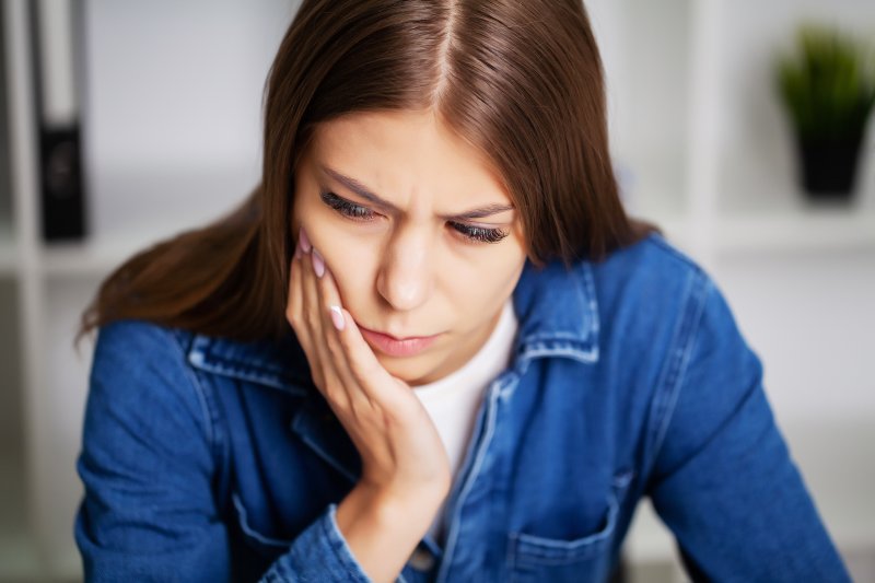 a young woman wearing a denim blouse and holding her cheek in pain because of a toothache