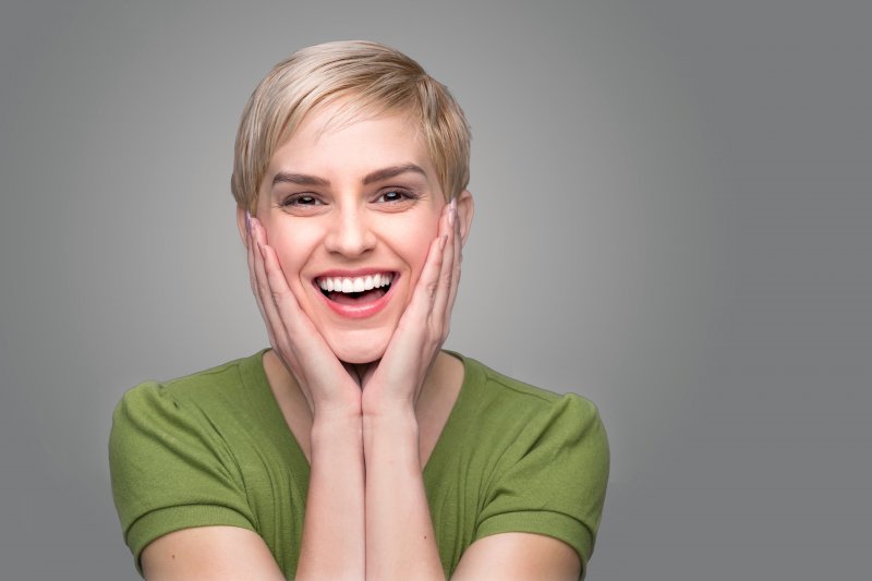 A young woman with blonde hair and wearing a green blouse holding her face between her hands and smiling 